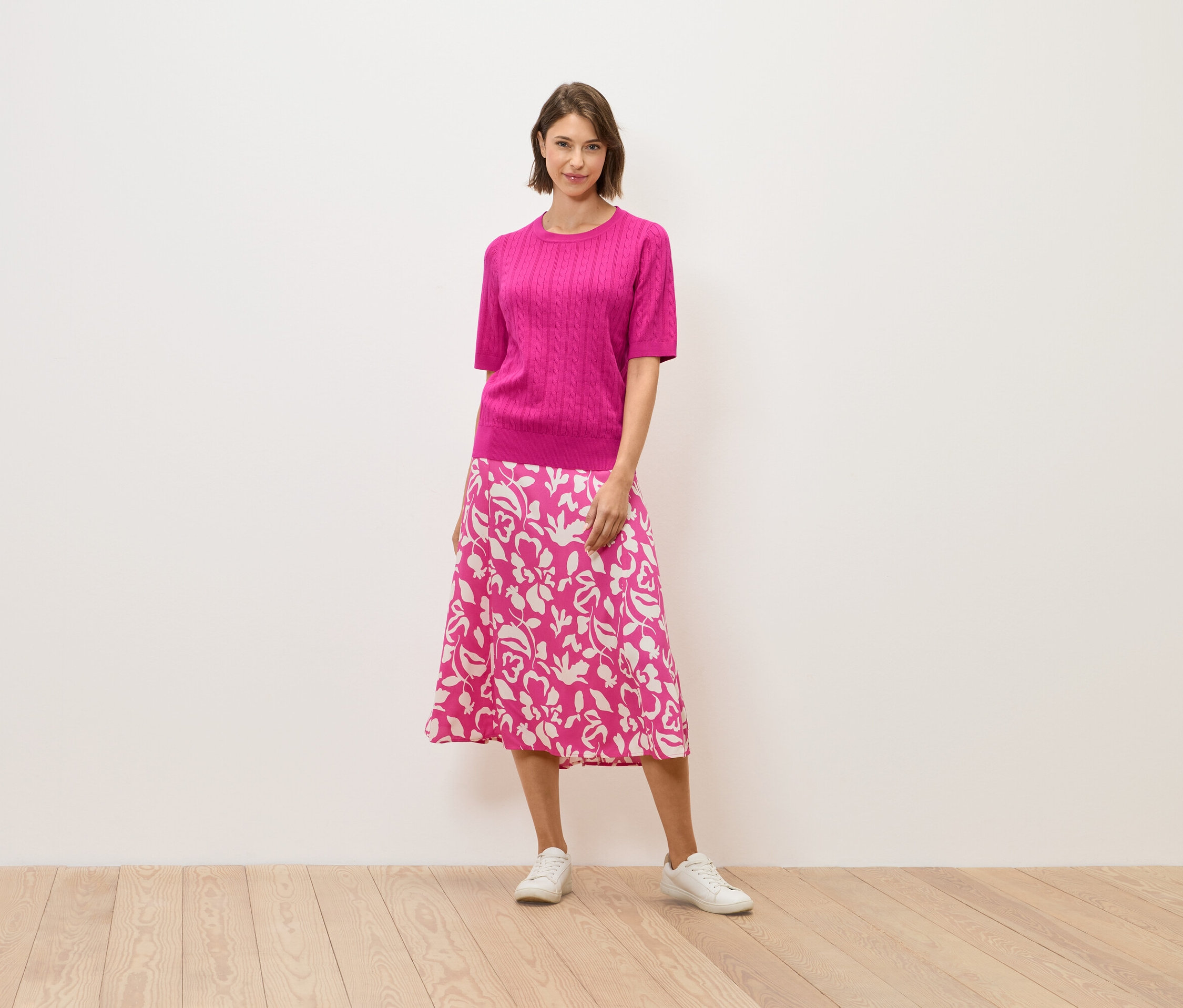 Une femme en pull rose et jupe à fleurs se tient sur un plancher en bois.