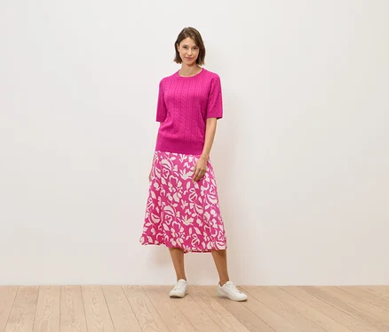 Une femme en pull rose et jupe à fleurs se tient sur un plancher en bois.