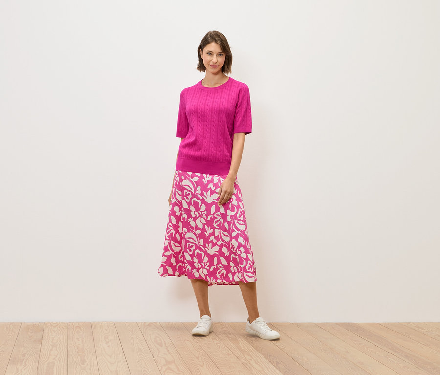 Une femme en pull rose et jupe à fleurs se tient sur un plancher en bois.
