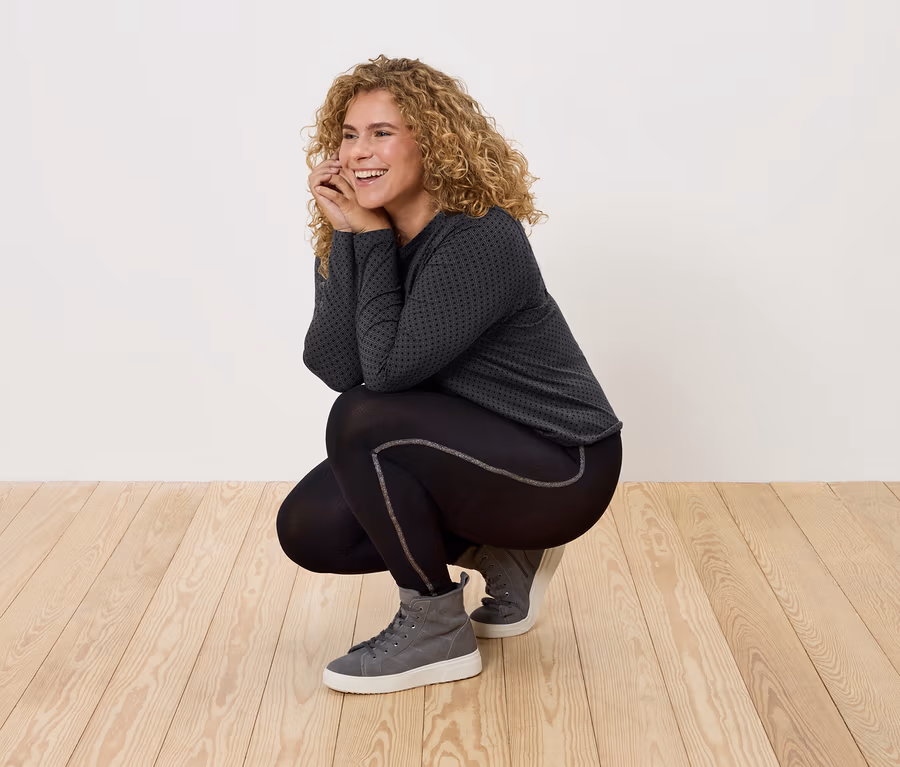 Une femme aux cheveux bouclés, vêtue de vêtements de sport noirs et de baskets grises, pose sur un plancher en bois.