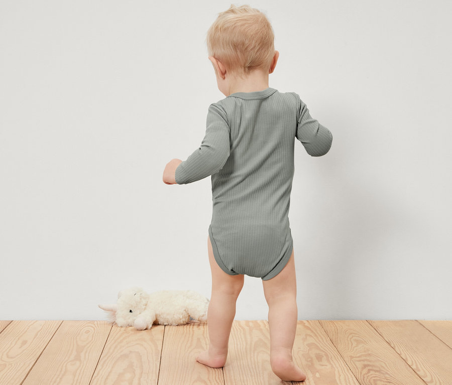 Un jeune enfant portant un body côtelé à manches longues se tient sur un plancher en bois à côté d'un animal en peluche.
