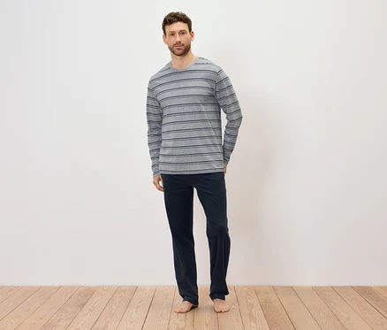 Un homme se tient pieds nus sur un plancher en bois, portant un t-shirt à manches longues gris rayé et un pantalon bleu marine.