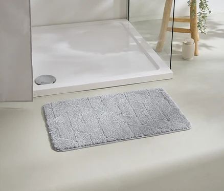 Un tapis de bain gris est posé devant un receveur de douche blanc, avec un tabouret et une plante en arrière-plan.