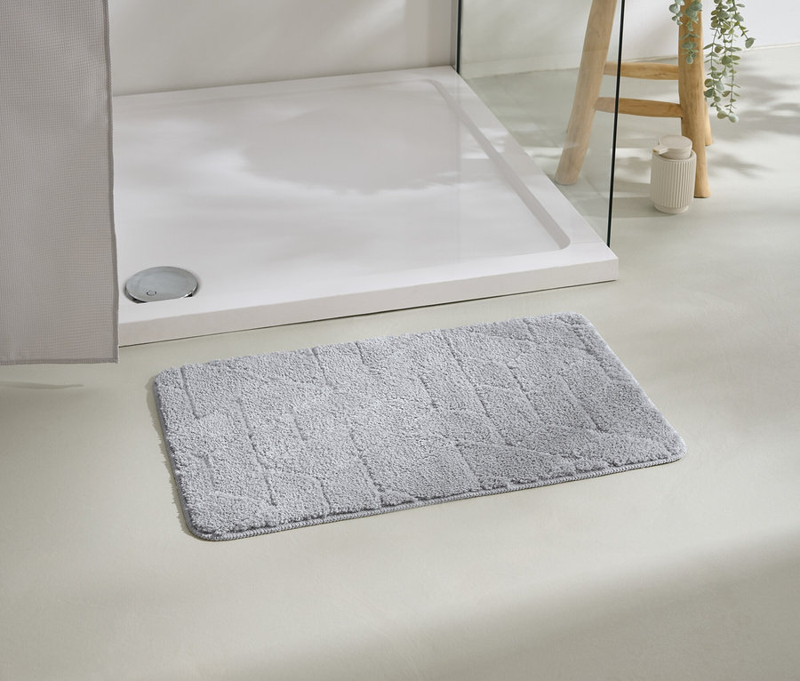 Un tapis de bain gris est posé devant un receveur de douche blanc, avec un tabouret et une plante en arrière-plan.