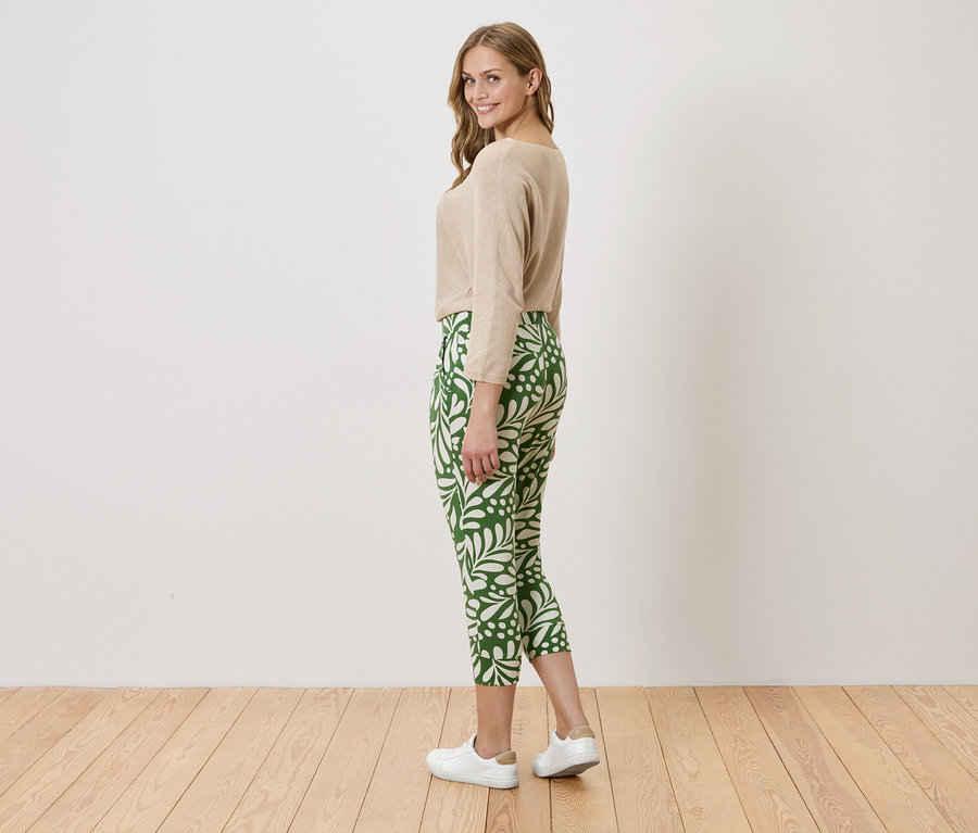 Femme portant un pull beige et un pantalon vert avec un motif blanc, debout sur un plancher en bois devant un mur blanc.