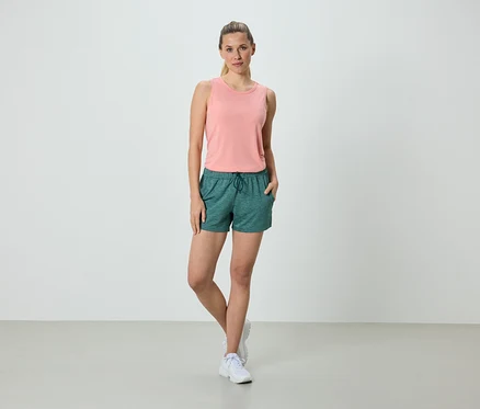 Femme debout avec les mains dans les poches en short vert et débardeur rose.