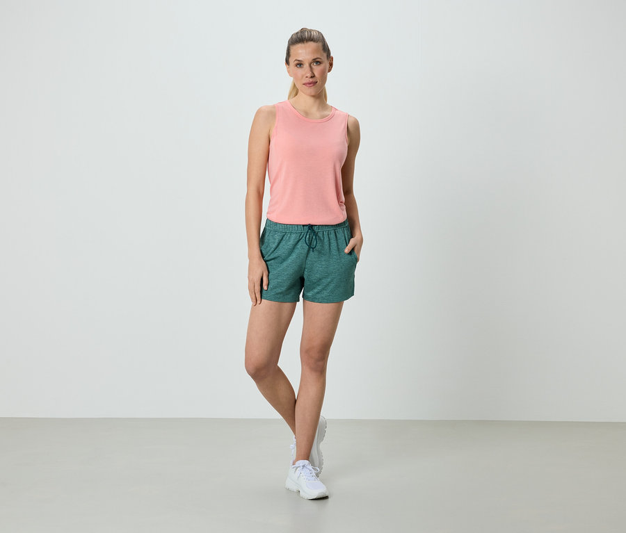 Femme debout avec les mains dans les poches en short vert et débardeur rose.
