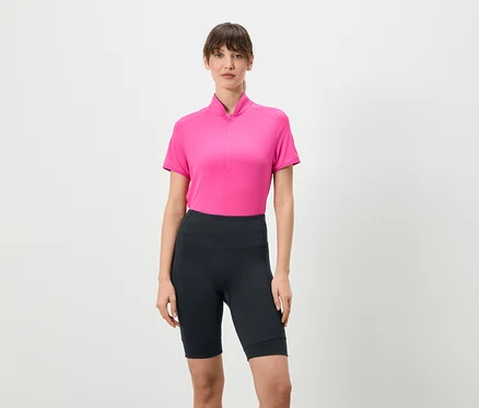 Une femme se tient debout sur un fond blanc, portant un t-shirt rose à fermeture éclair courte et des leggings courts foncés.