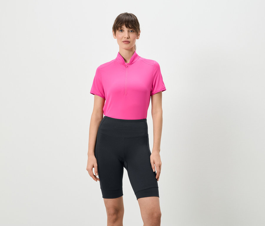 Une femme se tient debout sur un fond blanc, portant un t-shirt rose à fermeture éclair courte et des leggings courts foncés.