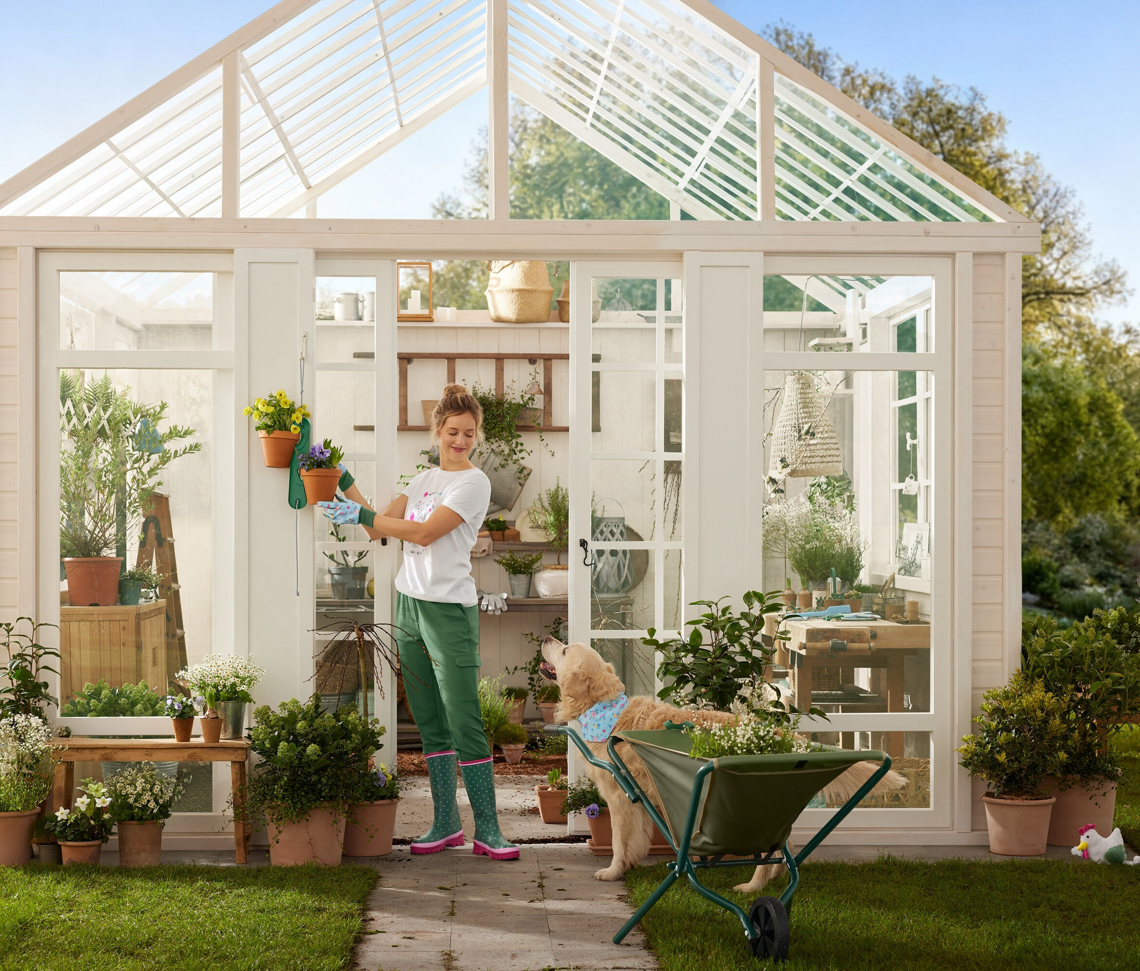 Une femme avec des gants de jardinage accroche des pots de fleurs à une serre, tandis qu'un chien se tient à côté d'une brouette.