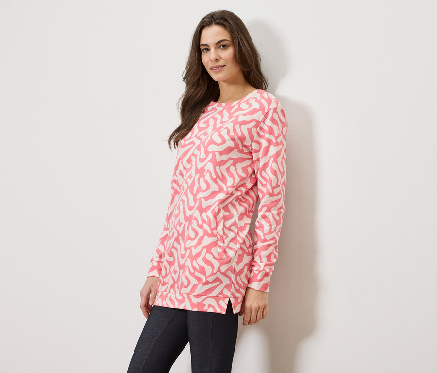 Une femme porte un pull rose à motifs abstraits et un jean foncé sur un fond blanc.