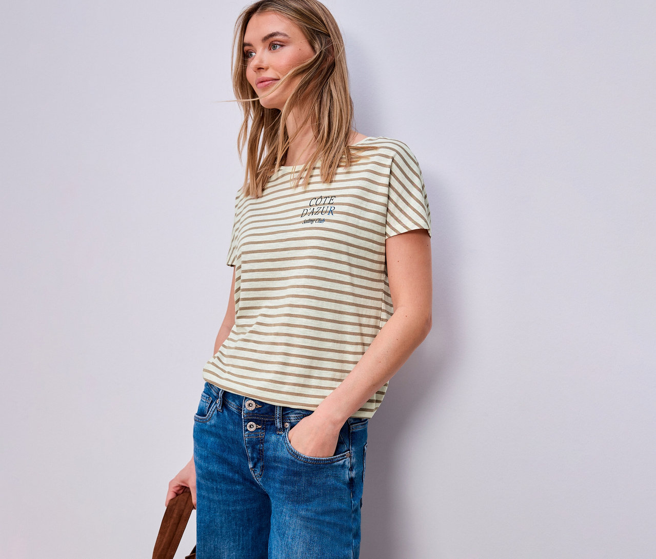 Femme portant un jean et un t-shirt Street One beige et blanc. Elle a une main dans sa poche et tient un sac marron.