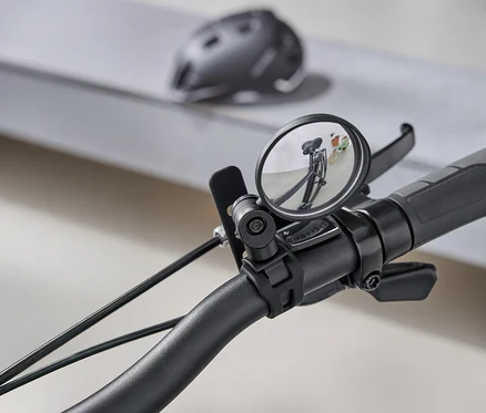 Gros plan sur un guidon de vélo avec un rétroviseur rond reflétant la route derrière le vélo. Un casque de vélo gris est visible en arrière-plan.