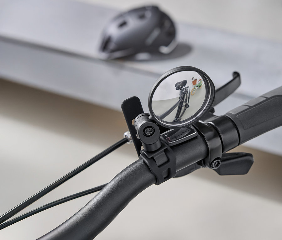 Gros plan sur un guidon de vélo avec un rétroviseur rond reflétant la route derrière le vélo. Un casque de vélo gris est visible en arrière-plan.