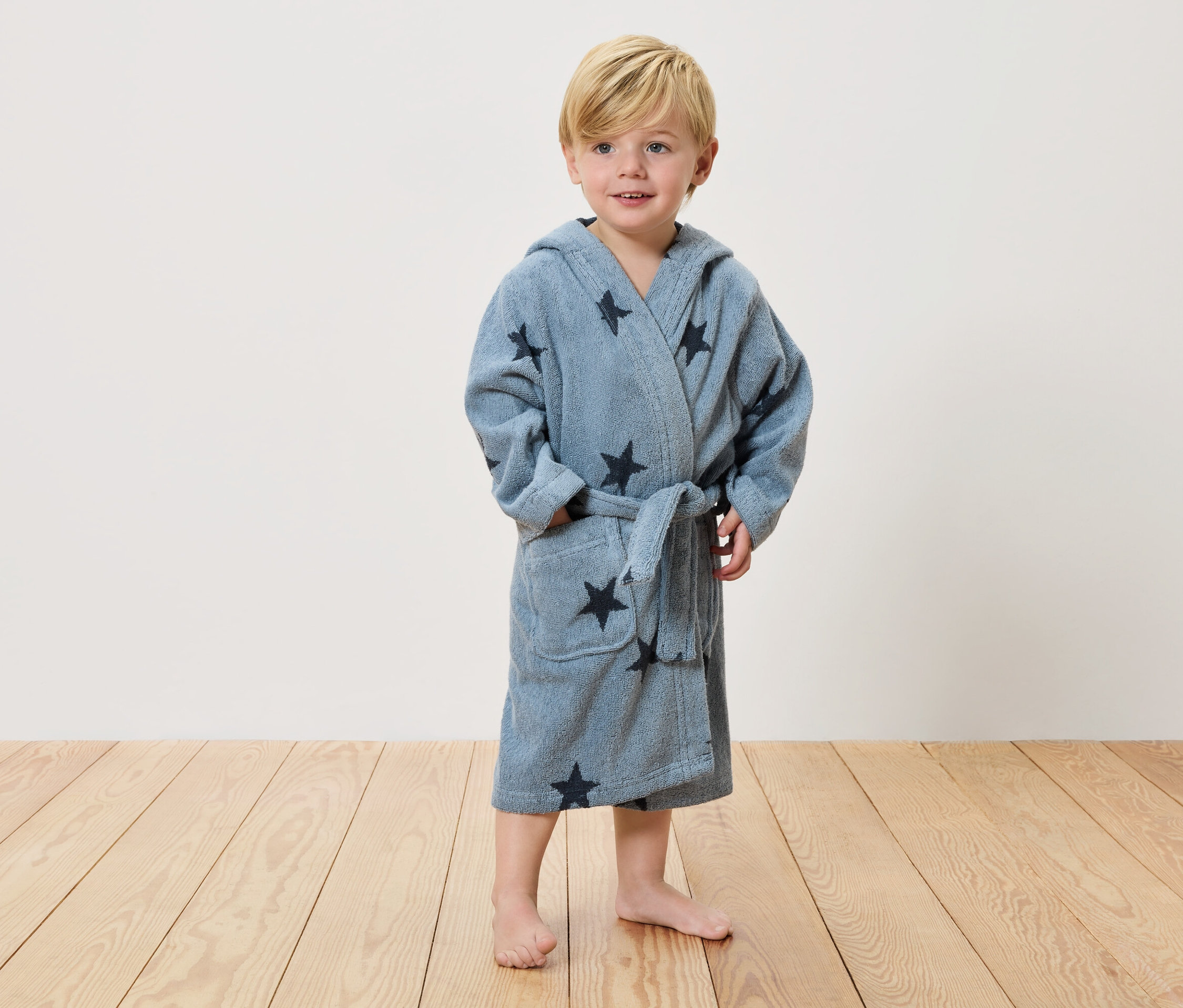 Un petit garçon blond se tient pieds nus sur un plancher en bois et porte un peignoir bleu clair avec des étoiles foncées.