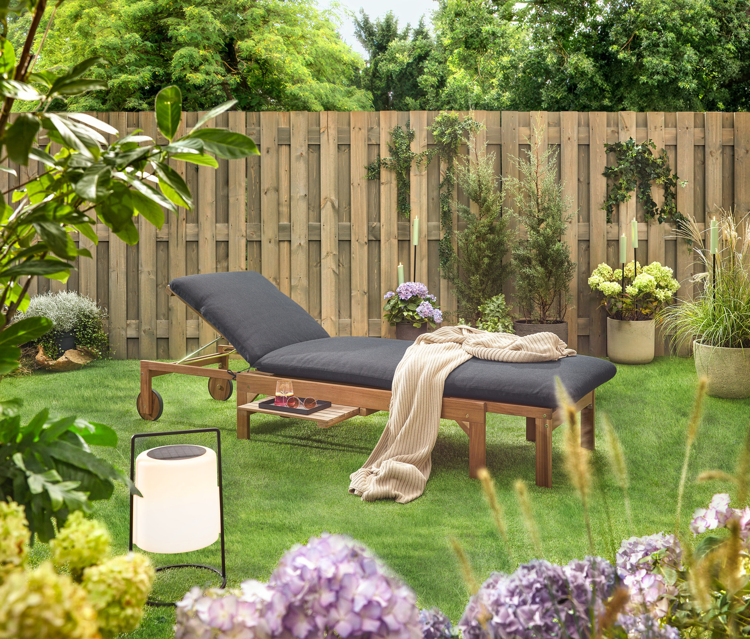 Scène de jardin avec une chaise longue en bois avec un rembourrage noir, une couverture, une boisson, des lunettes de soleil et une lanterne sur une pelouse devant une clôture en bois.