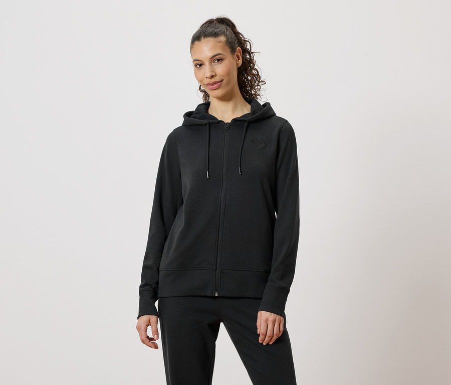 Une femme porte un survêtement noir composé d'un sweat à capuche zippé et d'un pantalon sur un fond neutre.