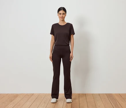 Une femme en t-shirt marron et pantalon évasé se tient devant un mur blanc sur un plancher en bois.