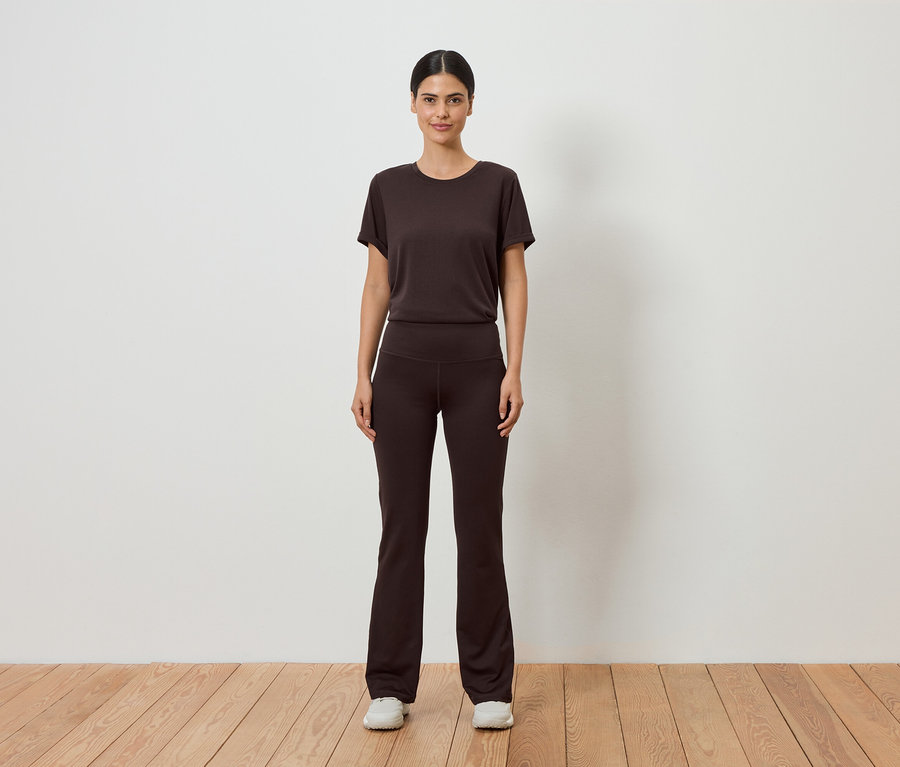 Une femme en t-shirt marron et pantalon évasé se tient devant un mur blanc sur un plancher en bois.