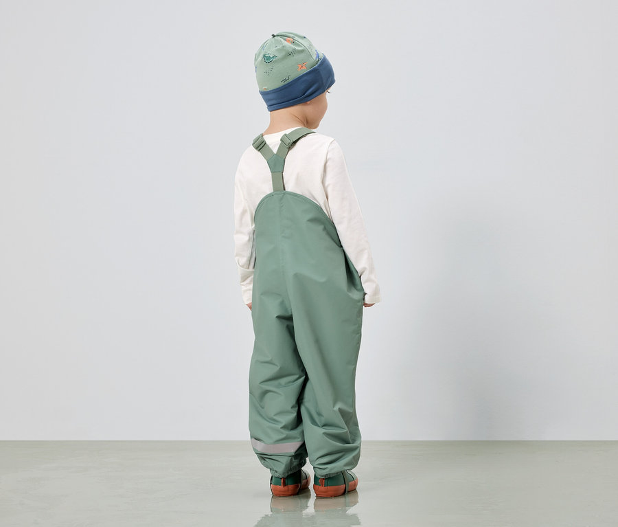Un enfant, vu de dos, porte une salopette verte, un haut blanc à manches longues et un bonnet vert à motifs de dinosaures.