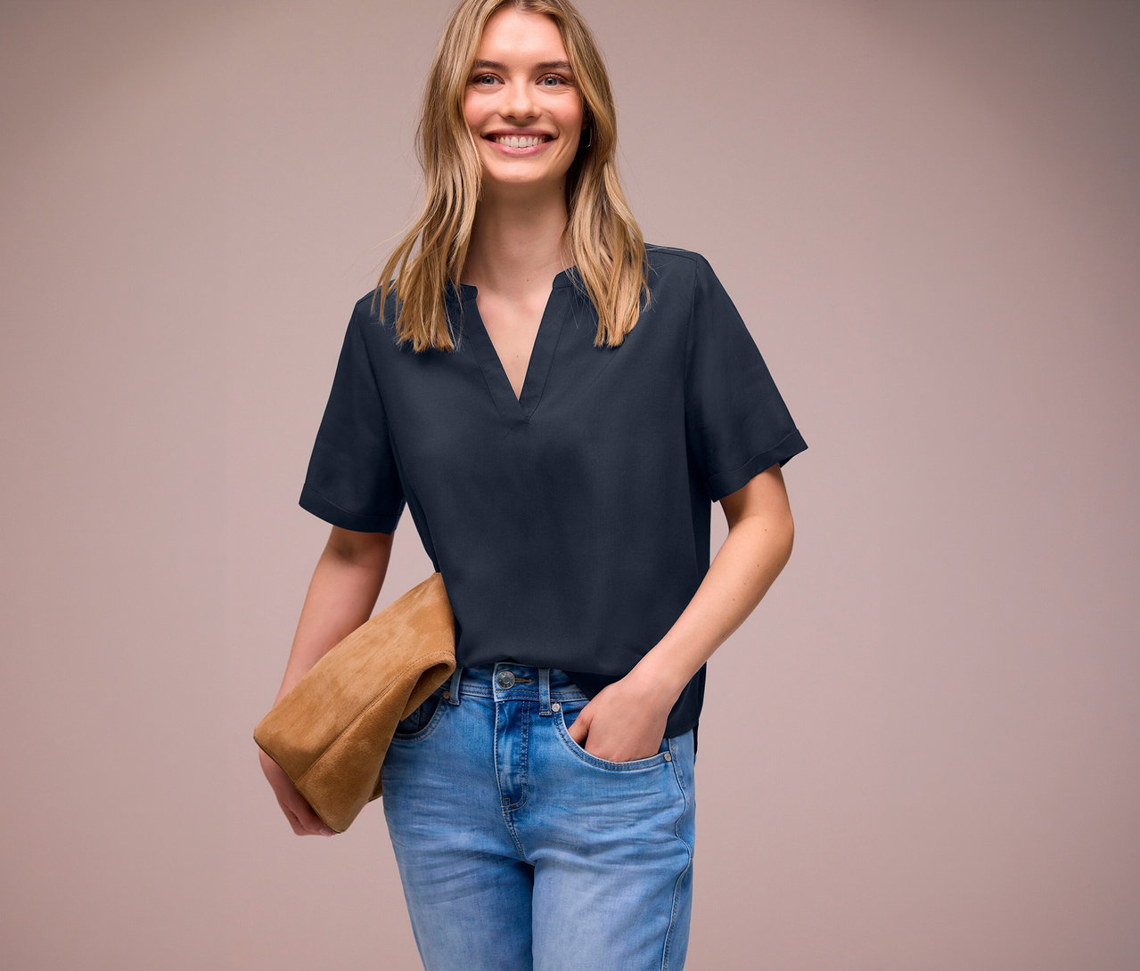 Une femme souriante porte un jean bleu et une blouse en maille Street One, et tient un sac en daim marron.