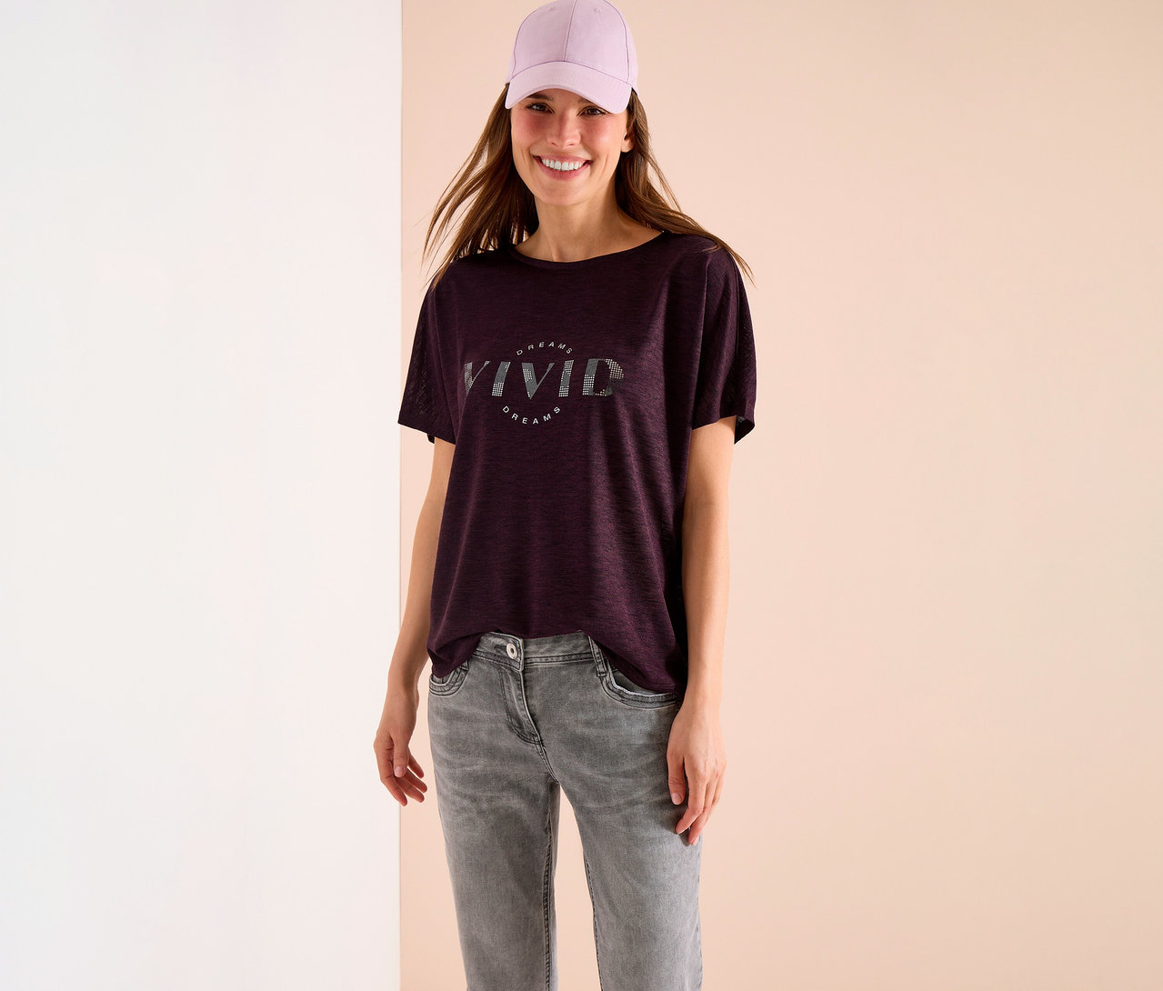 Une femme portant une casquette violette, un t-shirt bordeaux avec l'inscription "Vivid Dreams" et un jean gris.