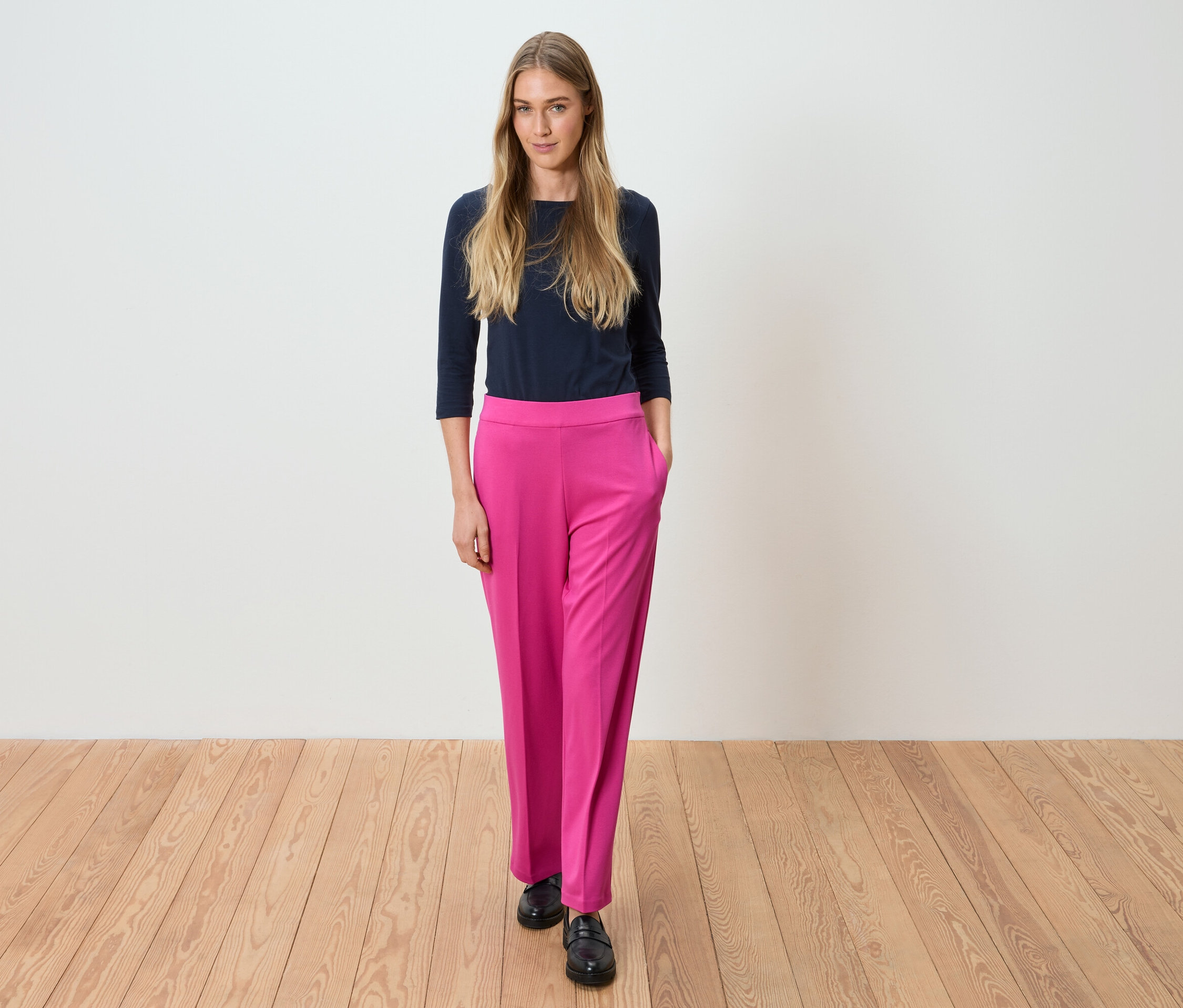 Une femme se tient devant un mur blanc, portant un haut bleu marine et un pantalon rose.