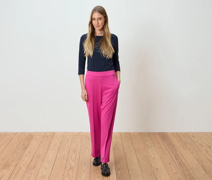 Une femme se tient devant un mur blanc, portant un haut bleu marine et un pantalon rose.