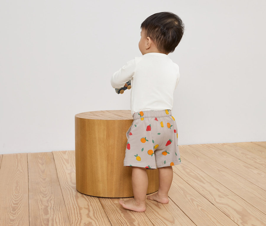 Un jeune enfant se tient dos au spectateur sur un plancher en bois, s'appuyant sur un tabouret en bois avec un jouet.