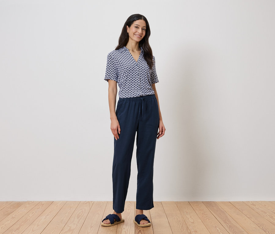 Une femme se tient devant un mur, portant une chemise géométrique bleue et un pantalon bleu marine.