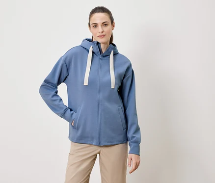 Une femme avec une queue de cheval porte un sweat à capuche bleu zippé et un pantalon beige.