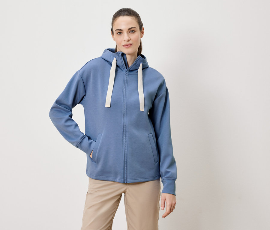 Une femme avec une queue de cheval porte un sweat à capuche bleu zippé et un pantalon beige.