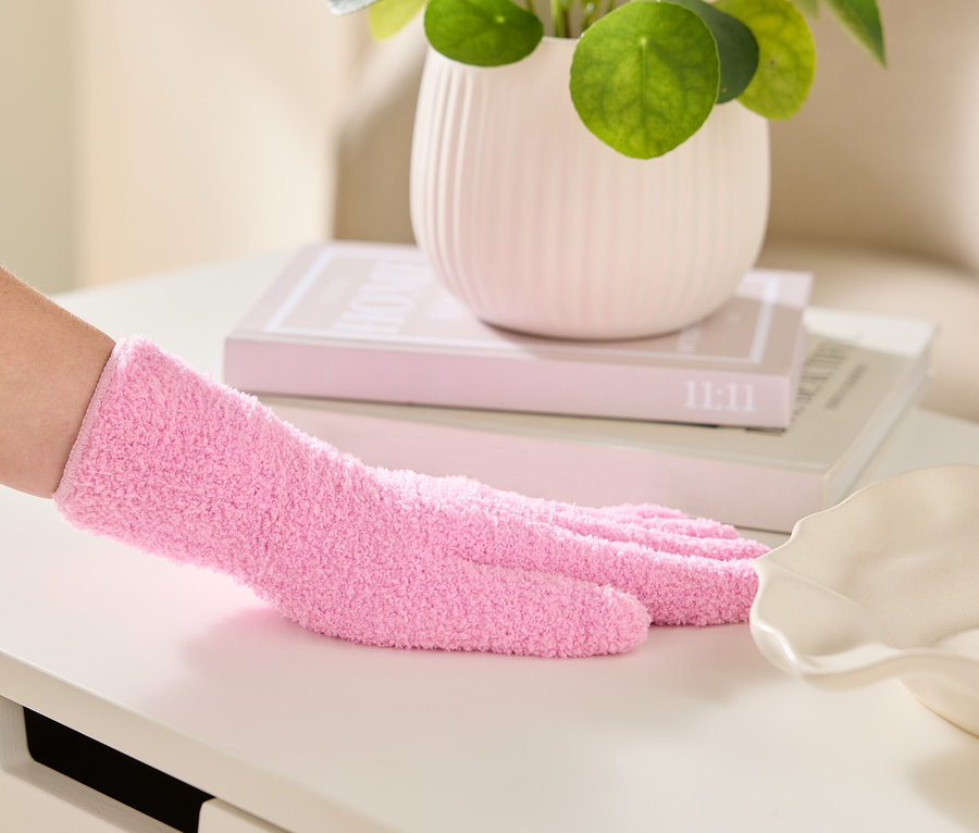 Une main gantée d'un gant en chenille rose essuie une surface blanche, avec une plante d'intérieur et des livres en arrière-plan.
