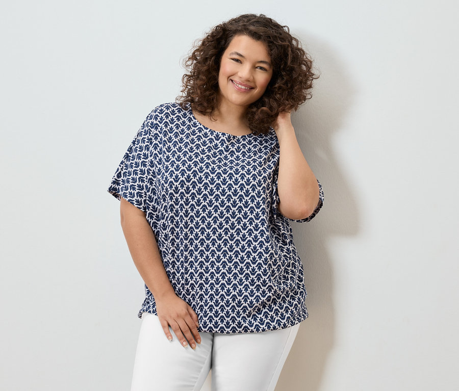 Femme souriante aux cheveux bouclés portant un chemisier bleu et blanc et un pantalon blanc.