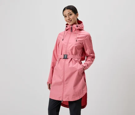 Une femme avec une capuche et une ceinture porte un imperméable rose et un pantalon noir devant un fond gris clair.