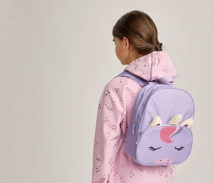 Une jeune personne vue de dos, portant une veste rose à motifs et un sac à dos licorne violet avec un visage, des oreilles et une corne de licorne.