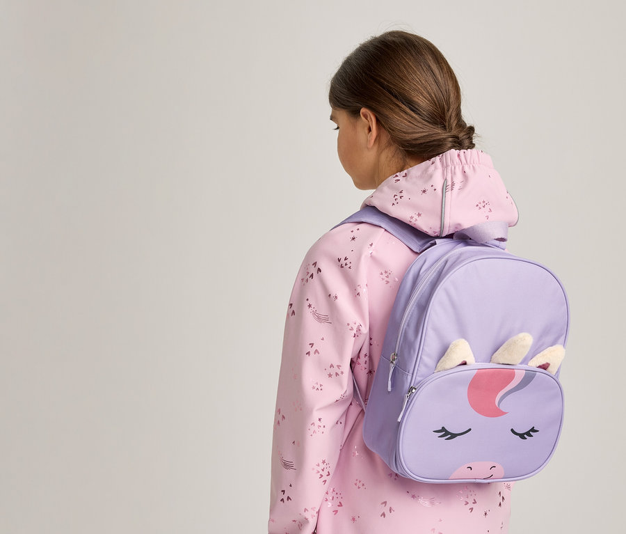 Une jeune personne vue de dos, portant une veste rose à motifs et un sac à dos licorne violet avec un visage, des oreilles et une corne de licorne.