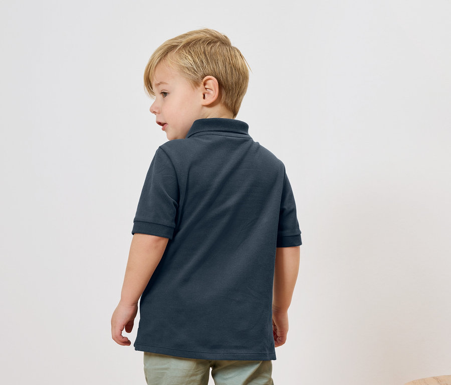 Un garçon blond porte un polo bleu marine et un pantalon vert clair, vu de dos.
