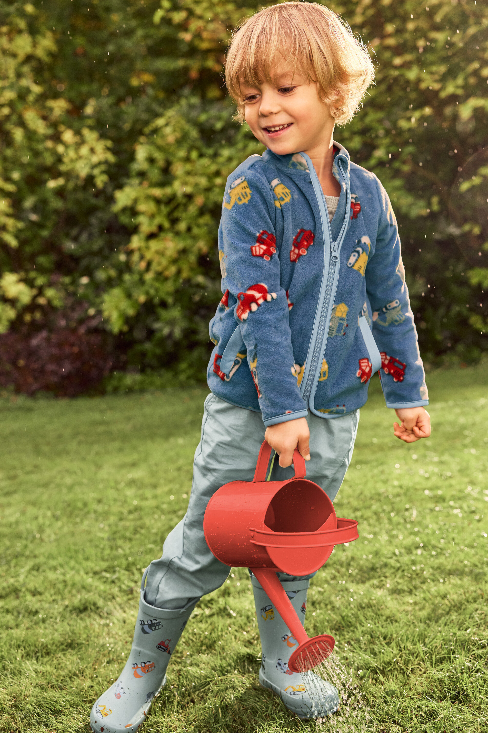 Un garçon blond souriant arrose une pelouse verte avec un arrosoir rouge. Il porte une veste bleue avec des motifs de véhicules et des bottes en caoutchouc grises avec des véhicules.