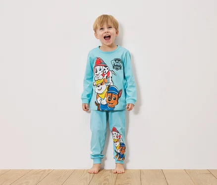 Un petit garçon se tient devant un mur blanc et porte un pyjama bleu clair Pat' Patrouille.