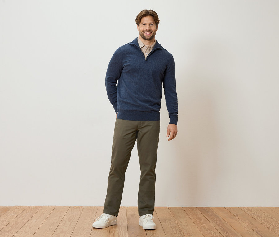 Un homme se tient devant un mur, les mains dans les poches. Il porte un pull bleu marine, un chemisier marron clair, un pantalon kaki et des baskets blanches.