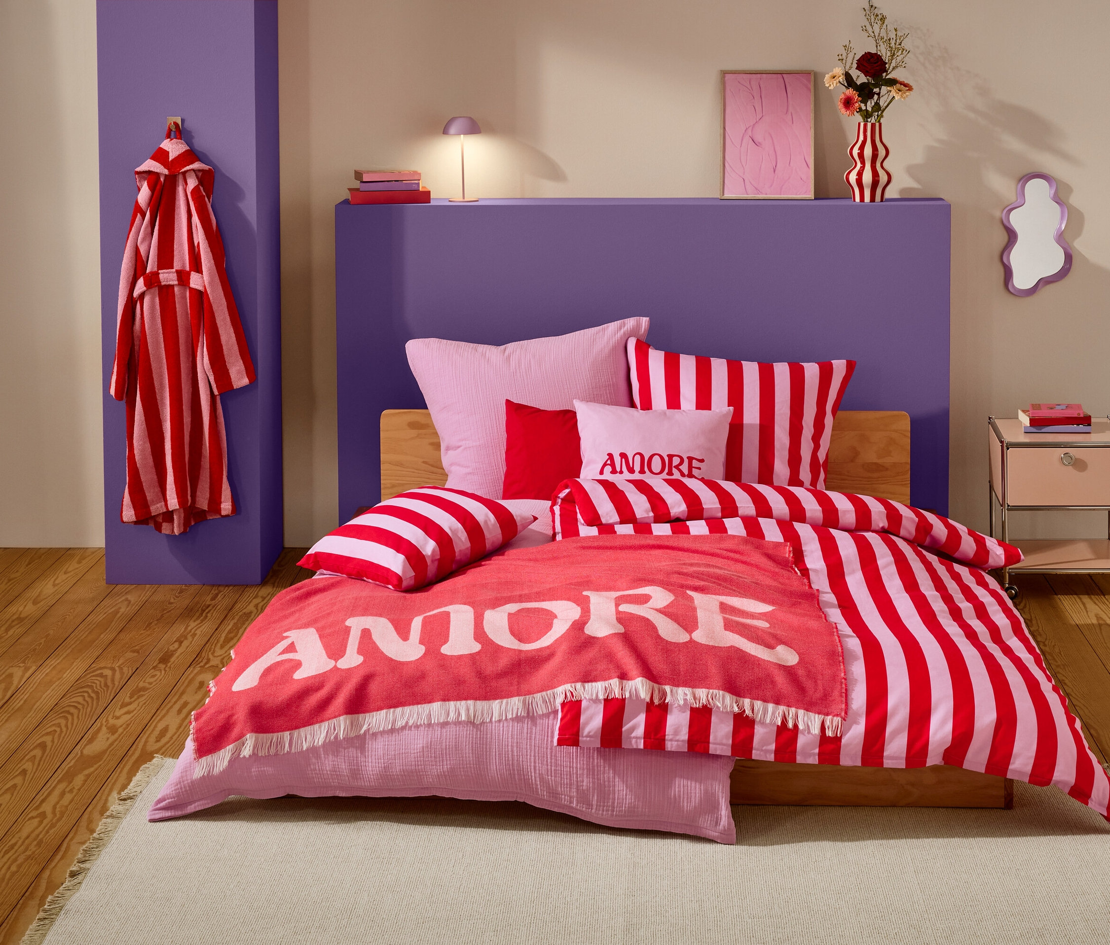 Une chambre avec un lit recouvert de linge de lit à rayures rouges et blanches, des oreillers roses et une couverture corail avec l'inscription "Amore". Un peignoir rayé rouge et blanc est accroché à un pilier violet.