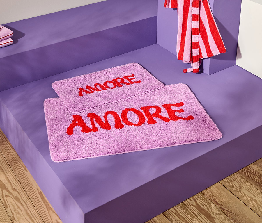 Photo de deux tapis de bain lavande avec le mot rouge "AMORE" sur une surface violette, ainsi que des serviettes rayées.