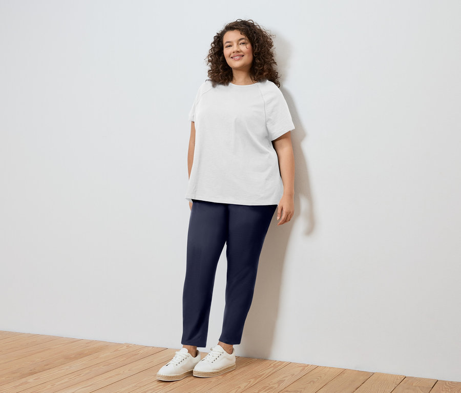 Une femme aux cheveux bouclés, portant un t-shirt blanc, un pantalon bleu et des chaussures blanches, se tient contre un mur blanc.