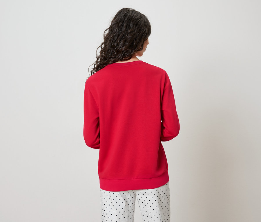 Vue de dos d'une femme aux longs cheveux ondulés et foncés, portant un pull rouge et un pantalon à pois.