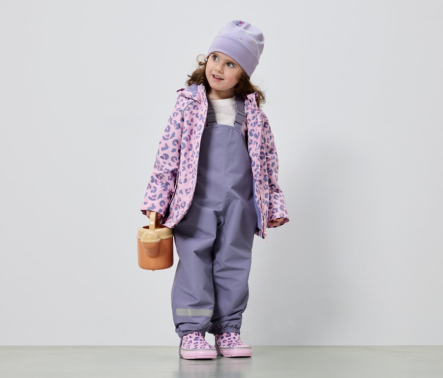 Une fillette aux cheveux bouclés porte une salopette violette, une veste rose et violette à motif léopard, un bonnet et des chaussures assorties. Elle tient un arrosoir.