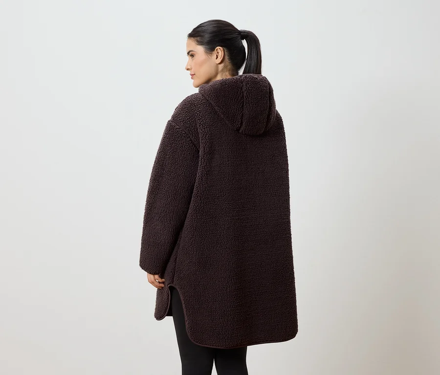 Une femme vue de dos, avec une queue de cheval, porte un sweat à capuche marron ample en tissu type teddy bear sur un fond blanc.