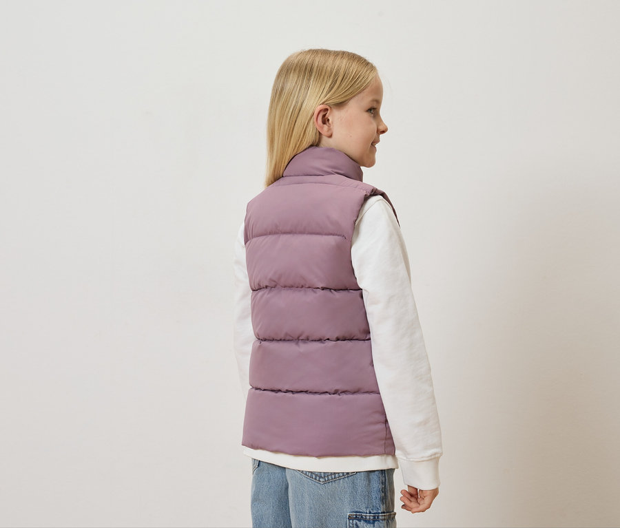 Une fille blonde porte un gilet matelassé mauve sur un pull blanc et un jean.