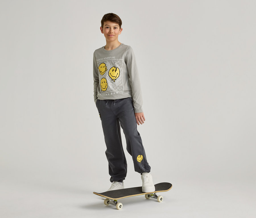 Un garçon portant un sweat-shirt et un pantalon gris avec des smileys jaunes se tient sur un skateboard devant un fond blanc.