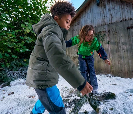 Deux enfants jouent dans la neige. Un enfant au premier plan porte une doudoune verte et un pantalon bleu et noir. Un autre enfant à l'arrière-plan équilibre sur une bûche enneigée, vêtu d'une veste verte et d'un pantalon de neige bleu foncé.